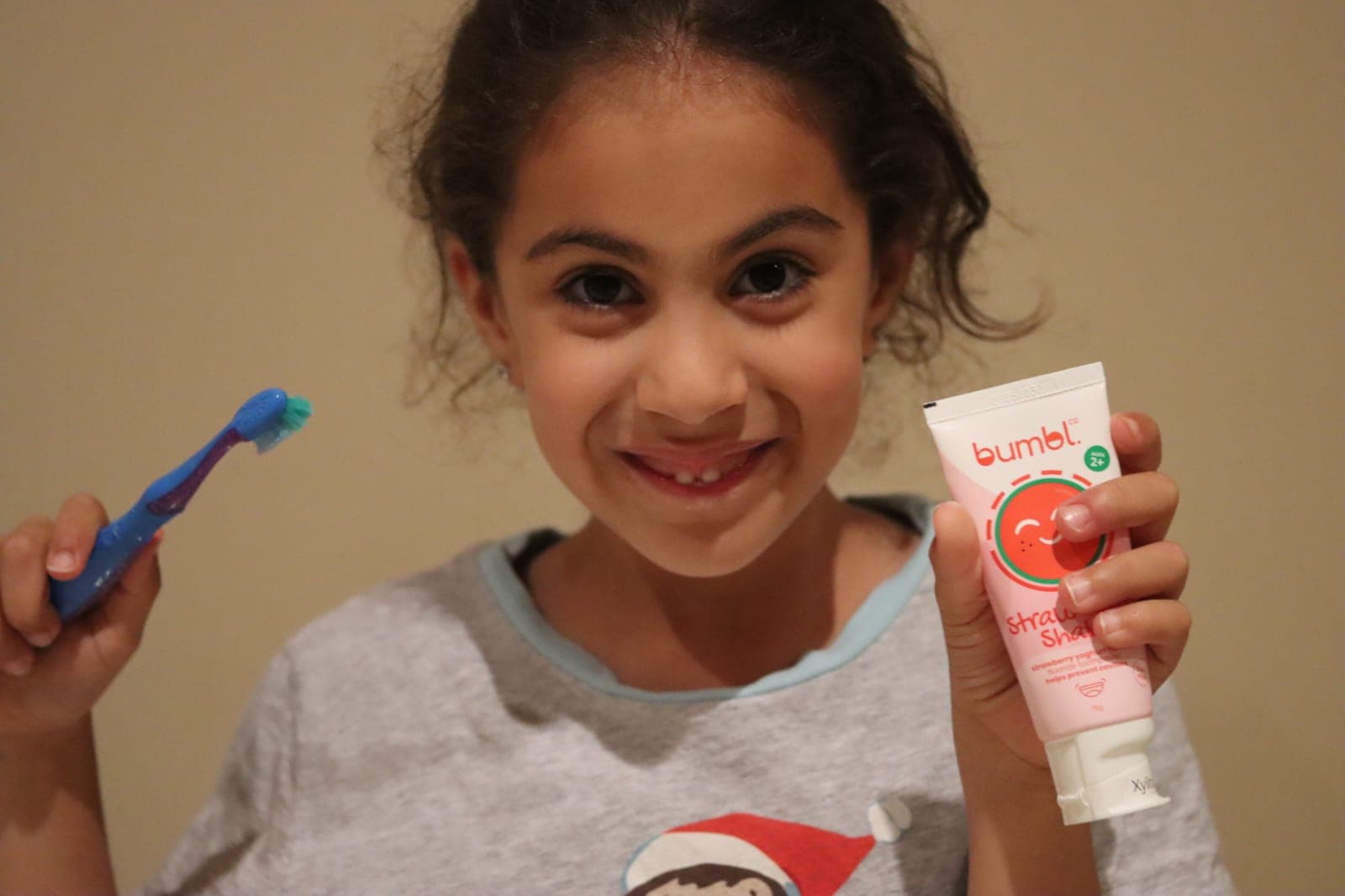 A smiling girl holding her toothbrush and a Strawberry Shake tube of Bumbl Co toothpaste
